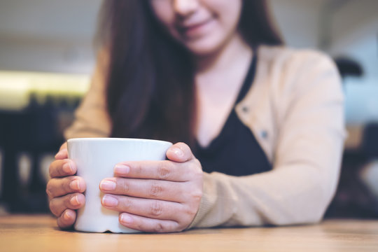 Closeup image of a woman holding a white cup of coffee with two hands in modern cafe