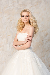 Fototapeta na wymiar fashionable white gown, beautiful blonde model, bride hairstyle and makeup concept - young smiling girl in wedding festive dress, standing indoors on light background, slender woman look forward