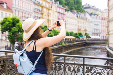 Fototapeta premium Woman taking pictures with smartphone. Stylish summer traveler woman in hat with camera outdoors in european city, old town Karlovy Vary in the background, Czech Republic, Europe