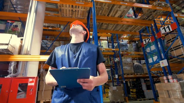A man tries to see high stored items on a warehouse and takes notes. 4K.