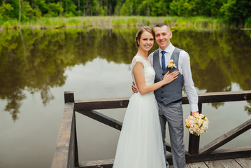 Young couple in love. Groom and bride in wedding dress gainst a wooden verandah on the lake watching to camera. Wedding. Wedding day. Bridal bouquet.