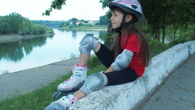 A beautiful girl in roller skates sits on the river bank. A little girl on roller skates is at the river.
