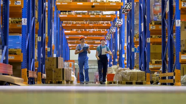 Two warehouse workers walk under high orange storage racks.