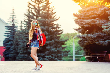 Fashion portrait of young pretty hipster woman outdoor with long hair and red backpack in the sunny summer street. The evening sunset over the city.