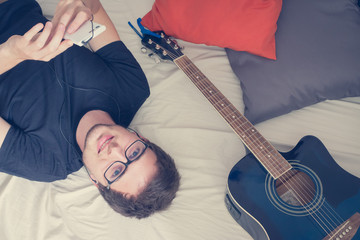 Junger Mann liegend im Bett, Gitarre und Smartphone, sorgenlos