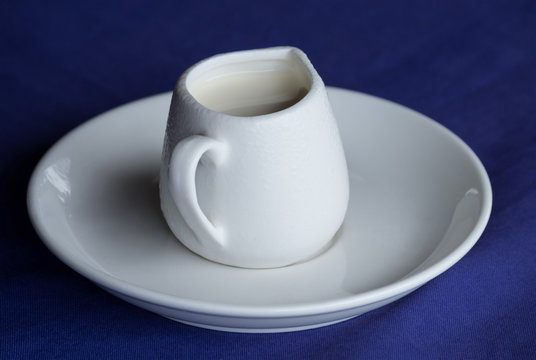 White ceramic creamer on a saucer with cold cream on a blue background.