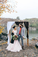 Stylish bride and groom with dreadlocks are holding hands next to wedding arch. Autumn wedding...