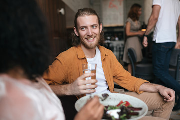 Smiling man sitting in restaurant and talking with girl. Boy sitting at cafe with glass of water in hand. Young joyful man with blond hair and beard happily looking at girl in restaurant