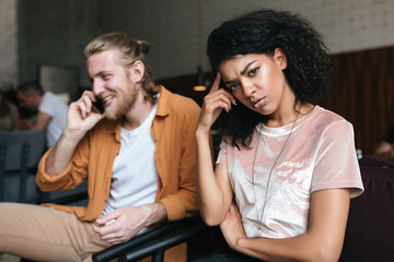 Portrait of young man and girl sitting in restaurant. Upset African American girl with dark curly hair sadly looking in camera while boy with blond hair happily talking on his cellphone at cafe