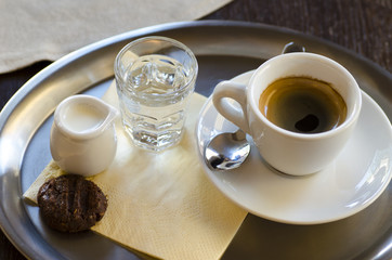 Close-up view of a cup of coffee with milk, a glass of water and a cracker standing on a stainless steel tray on a napkin