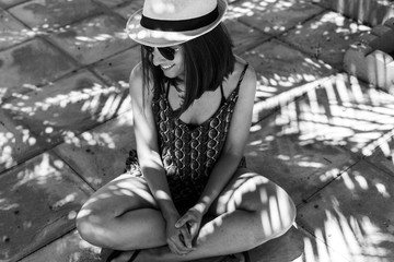Obraz na płótnie Canvas black and white portrait of a young beautiful woman wearing summer outfit. Sitting on the floor. Sunglasses and hipster hat. Sunny. Lifestyle
