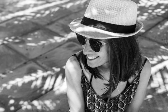 black and white portrait of a young beautiful woman wearing summer outfit. Sitting on the floor. Sunglasses and hipster hat. Sunny. Lifestyle