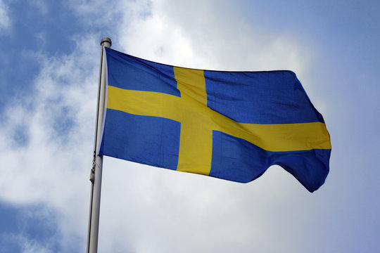 Flag of Sweden with a yellow cross on a blue background, national symbol or sign of the european country, fluttering in the wind against the blue sky with clouds on a sunny day