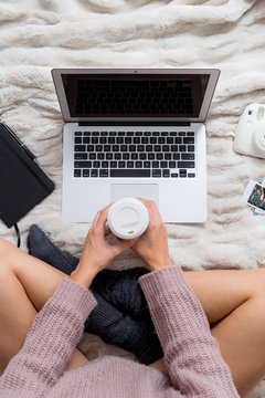 Overhead view of a woman in bed using a laptop while dinking coffee