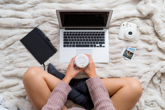 Overhead view of a woman in bed using a laptop while dinking coffee