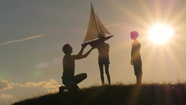 The father shows the model of the sailboat to the children. Backlight. Silhouettes of people against the sky and sun. Model of a sailboat.