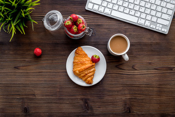Dessert fo light lunch at workplace. Coffee, strawberry, croissant near keyboard on wooden background top view copyspace