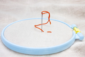 The beginning of needlework on an embroidery hoop. Needle and orange thread.
