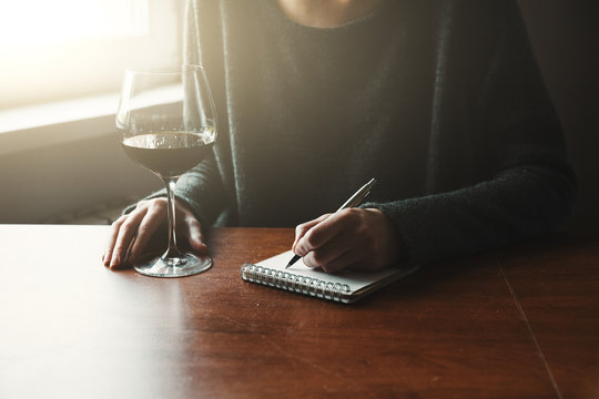 female hands with pen and glass of wine writing on notebook