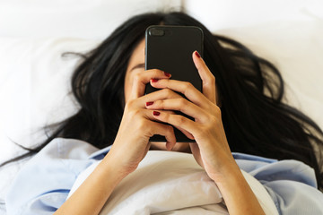 Top view of a beautiful young Asian woman holding a smart phone lying on the white bed. Focus close up hands using a smart phone.