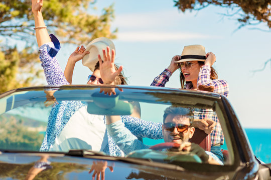 Group of happy young friends in cabriolet with raised hands driving on sunset