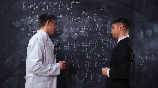 Two young successful modern scientists in formal suit, glasses and medical gown writing, discussing and analyzing math equations on a chalk desk board. Middle static shot.