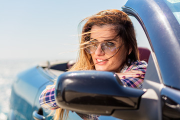 Front view of woman sitting in cabriolet putting hand on door looking away on background of sea.