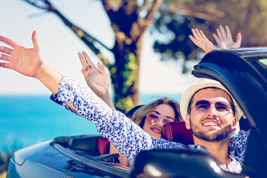 Group of happy young friends in cabriolet with raised hands driving on sunset