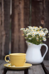 A yellow cup of chamomile tea with a pitcher of chamomile flowers in the background.