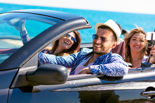 Group of happy young friends in cabriolet with raised hands driving on sunset