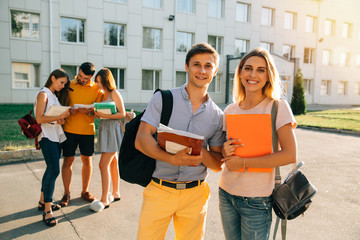 Two happy students with backpacks and books in their hands smiling at camera while standing on background of university and friends