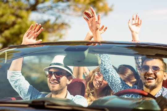 Group of happy young friends in cabriolet with raised hands driving on sunset