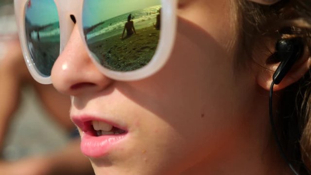 Teenage girl in sunglasses. Sits on the beach and sings a song listening to her in headphones with a smartphone. In the glasses reflects the beach with sea waves. 4k. Slow motion