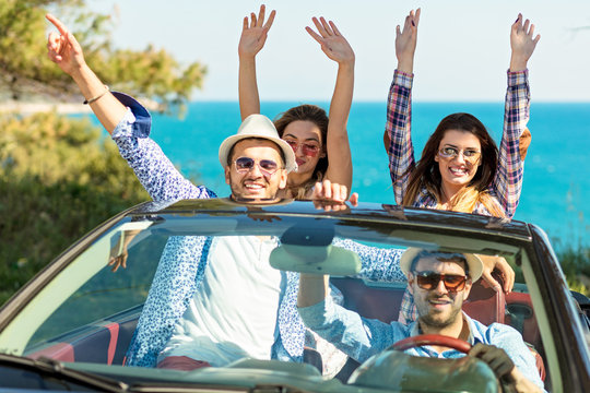 Group of happy young friends in cabriolet with raised hands driving on sunset