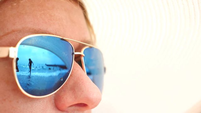 Close-up face of a happy woman in sunglasses. The beach with the resting people is reflected in the glasses. 4k, slow motion