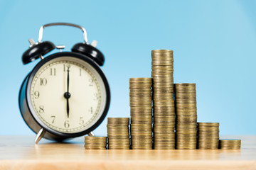 coins stack with alarm clock on wooden table and light blue background