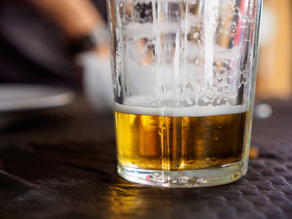 Close-up detail of the bottom of a near empty glass of beer, with foam on the sides, and a defocused person in the background. Valencia, Spain. Travel and drinks concept.