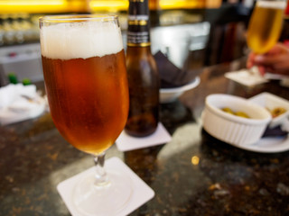 Close-up detail of an ale beer in a stemmed glass on a bar counter, with olives in ramekins as an appetizer, and a hand and glass in the background. Valencia, Spain. Travel and drinks concept.