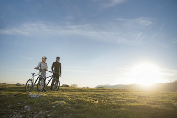 Fototapeta premium Cyclist couple with mountain bikes standing on the hill under the evening sky and enjoying bright sun at the sunset.