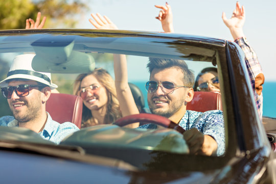 Group of happy young friends in cabriolet with raised hands driving on sunset
