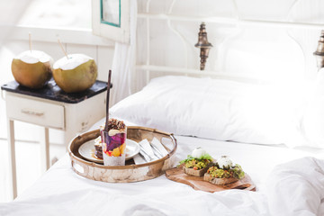 Fresh tasty breakfast in bed on wooden tray. Yogurt with fruits and chia, honey pancakes, two toasts with avocado and poached eggs and two young coconut. White vintage interior