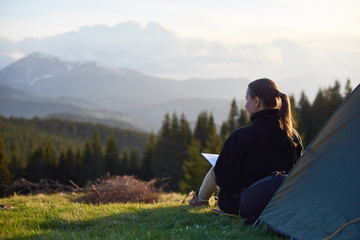 Fototapeta premium Rear view of young woman enjoying sunset in the mountains with a book near the tent