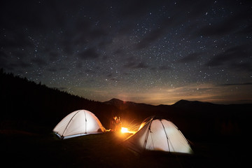 Night camping. Couple tourists sitting at a campfire near two illuminated tents under night starry sky. Long exposure