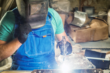 A young man in a welding mask in a blue construction overalls welds metal with a welding machine on a wooden table in the workshop, blue sparks from welding