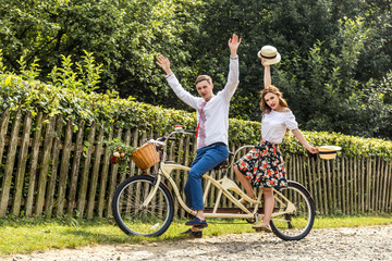 Young couple with bike tandem in park. Young people keep hats in their hands and smile. On the back...