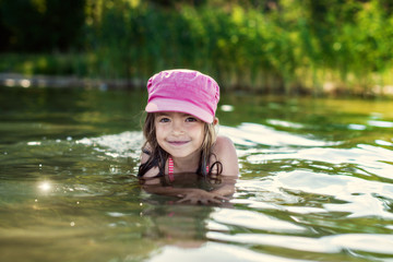 Smiling kid girl in pink cap having fun time and swimming in lake water. Concept of summer vacation