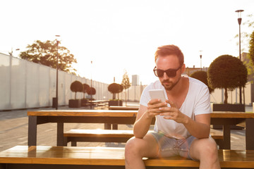 Modern guy sitting on the bench and using cellphone outdoors.