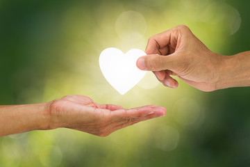 Hand holding and giving white heart to receiving hand on blurred green bokeh background, helping...