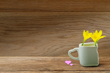 Fresh yellow flowers in white cup with heart shaped holder and pink heart on grunge wooden tabletop on vintage wooden wall