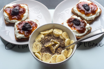 White bowl of cornflakes slice of banana and chia seeds, dark bread with white cheese and jam in background